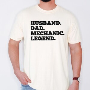 Puede incluir: Una camiseta blanca con texto negro que dice "Husband. Dad. Mechanic. Legend."