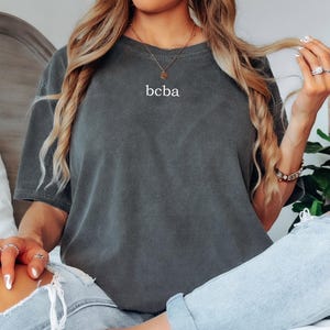 Op de afbeelding: Een vrouw draagt een donkergrijs oversized t-shirt met de tekst "bcba" erop gedrukt. Ze draagt een blauwe jeans en heeft haar haar naar achteren getrokken.