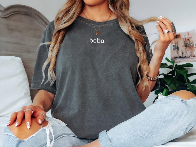 Op de afbeelding: Een vrouw draagt een donkergrijs oversized t-shirt met de tekst "bcba" erop gedrukt. Ze draagt een blauwe jeans en heeft haar haar naar achteren getrokken.