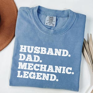 Puede incluir: Una camiseta azul claro con el texto "Husband. Dad. Mechanic. Legend." impreso en blanco.