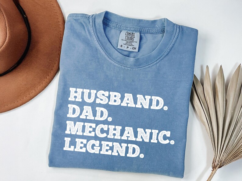 Puede incluir: Una camiseta azul claro con el texto "Husband. Dad. Mechanic. Legend." impreso en blanco.