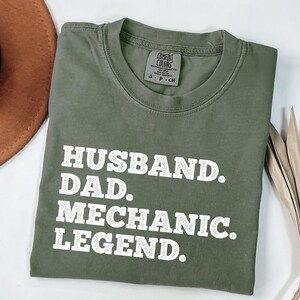 Puede incluir: Una camiseta verde con texto blanco que dice "Husband. Dad. Mechanic. Legend."