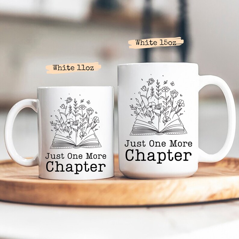 Puede incluir: Dos tazas de cer&aacute;mica blanca con un contorno negro de un libro abierto con flores que salen de &eacute;l. El texto en las tazas dice "Just One More Chapter". Las tazas est&aacute;n sobre una superficie de madera.