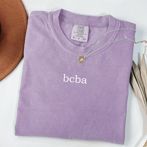 Op de afbeelding: Een lavendelkleurig T-shirt met de tekst "bcba" geborduurd op de voorkant. Het shirt is gemaakt door Comfort Colors en is maat S-P-CH.