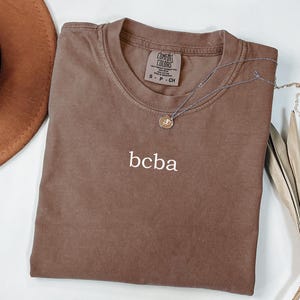 Op de afbeelding: Een bruin t-shirt met het woord "bcba" geborduurd op de voorkant. Het shirt is gemaakt van een zachte, comfortabele stof en heeft een ontspannen pasvorm.