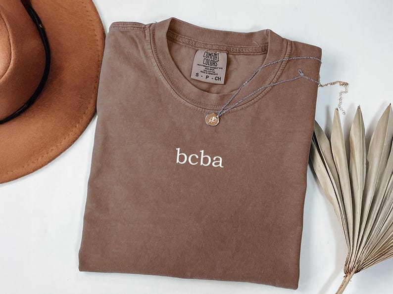 Op de afbeelding: Een bruin t-shirt met het woord "bcba" geborduurd op de voorkant. Het shirt is gemaakt van een zachte, comfortabele stof en heeft een ontspannen pasvorm.
