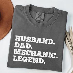 Puede incluir: Camiseta gris con texto blanco que dice "Husband. Dad. Mechanic. Legend."