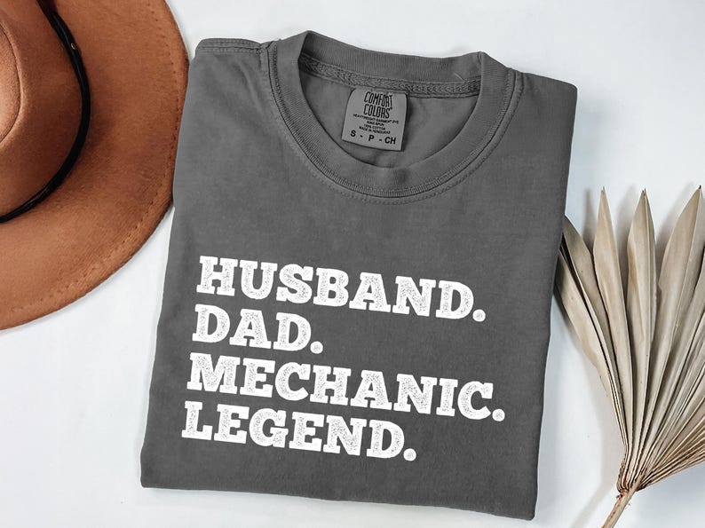 Puede incluir: Camiseta gris con texto blanco que dice "Husband. Dad. Mechanic. Legend."
