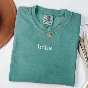 Op de afbeelding: Een tealblauw t-shirt met de tekst "bcba" geborduurd op de voorkant. Het shirt heeft een label met de tekst "Comfort Colors" en "S - P - CH".