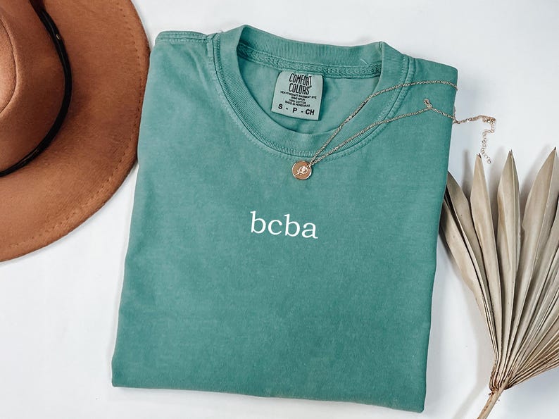 Op de afbeelding: Een tealblauw t-shirt met de tekst "bcba" geborduurd op de voorkant. Het shirt heeft een label met de tekst "Comfort Colors" en "S - P - CH".