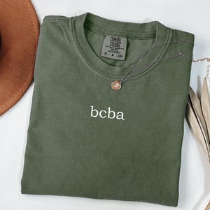 Op de afbeelding: Een groen T-shirt met de tekst "bcba" geborduurd op de voorkant. Het shirt is van Comfort Colors en is maat S - P - CH.