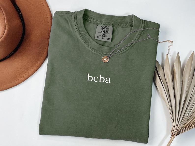 Op de afbeelding: Een groen T-shirt met de tekst "bcba" geborduurd op de voorkant. Het shirt is van Comfort Colors en is maat S - P - CH.