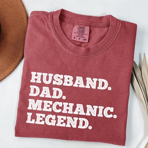 Puede incluir: Una camiseta de color marr&oacute;n rojizo con texto blanco que dice "Husband. Dad. Mechanic. Legend."