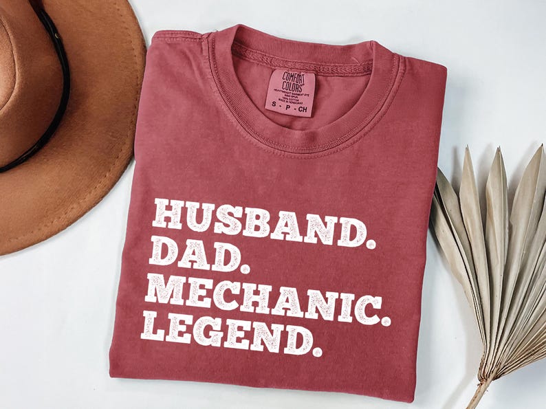 Puede incluir: Una camiseta de color marr&oacute;n rojizo con texto blanco que dice "Husband. Dad. Mechanic. Legend."