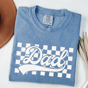 May include: A light blue t-shirt with a white checkered pattern and the word "Dad" in a retro font. The shirt has a crew neck and short sleeves.