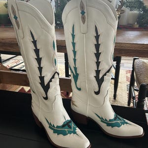 May include: A pair of white cowboy boots with intricate teal and black stitching. The boots have a pointed toe and a mid-height heel. The design includes decorative stitching along the shaft and vamp, creating a classic Western look.