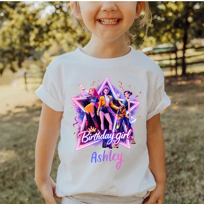 May include: White t-shirt featuring a colorful graphic of three animated figures inside a star-shaped frame. The text "Birthday girl" is above the name "Ashley" in a gradient font. The design includes pink, purple, yellow, and blue colors.