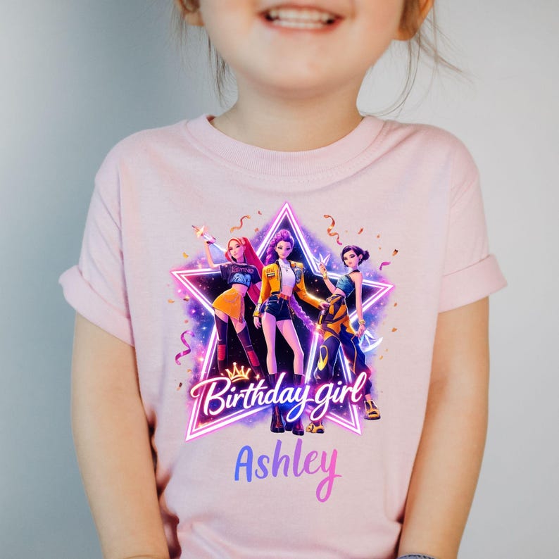 May include: Pink t-shirt featuring a graphic of three stylized figures inside a star, with the text "Birthday girl" and the name "Ashley" in a gradient font. The figures are in various outfits with vibrant colors.