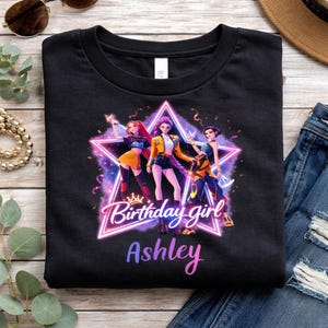 May include: Black t-shirt featuring a graphic of three animated figures inside a neon star. The text "Birthday girl Ashley" is displayed below the star in a gradient font. The design includes a crown and colorful accents.