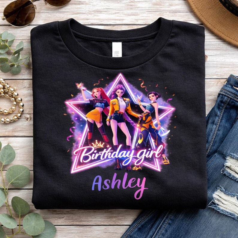 May include: Black t-shirt featuring a graphic of three animated figures inside a neon star. The text "Birthday girl Ashley" is displayed below the star in a gradient font. The design includes a crown and colorful accents.