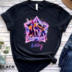 May include: Black t-shirt featuring a graphic of three animated figures inside a star-shaped frame. The text "Birthday girl" and the name "Ashley" are displayed in a gradient font. The shirt is made of a soft material.