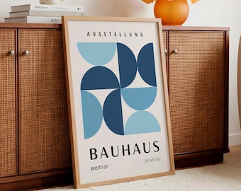 May include: A framed Bauhaus exhibition poster with a geometric design in shades of blue. The poster is propped against a wooden cabinet with woven doors. An orange glass lamp sits on top, and a mug and books are visible.