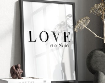 May include: Black framed print with the text "LOVE is in the air" on a white background. The print is on a black shelf with a gray vase and dried flowers.