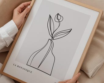 May include: A framed art print featuring a minimalist line drawing of a flower in a vase. The artwork is in black against a light background, with the words "LA BOTANIQUE" at the bottom. The frame is made of light-colored wood.
