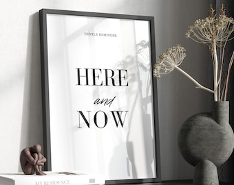 May include: Black framed poster with the text "GENTLE REMINDER HERE and NOW" on a white background. The poster is sitting on a black shelf with a small brown sculpture and a gray vase with dried flowers.