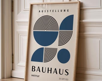 Op de afbeelding: Een ingelijste Bauhaus-tentoonstellingsposter. De poster heeft geometrische vormen in blauw en zwart op een crèmekleurige achtergrond. Het woord "AUSSTELLUNG" staat bovenaan, met "BAUHAUS" en "weimar" eronder. De data "JULI-SEPT.1923" staan onderaan.