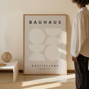 May include: A framed Bauhaus exhibition poster with a minimalist design of white geometric shapes on a light gray background. The poster includes the text "BAUHAUS" at the top and "AUSSTELLUNG JULI-SEPT 1923" at the bottom. The frame is a light brown color.