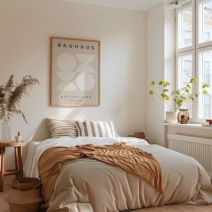 May include: A bedroom scene with a bed covered in a beige comforter and a brown throw blanket. Two striped pillows rest against the headboard. A framed Bauhaus poster hangs on the wall. A small wooden stool holds a vase with dried plants.