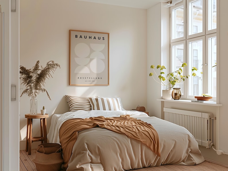 May include: A bedroom scene with a bed covered in a beige comforter and a brown throw blanket. Two striped pillows rest against the headboard. A framed Bauhaus poster hangs on the wall. A small wooden stool holds a vase with dried plants.