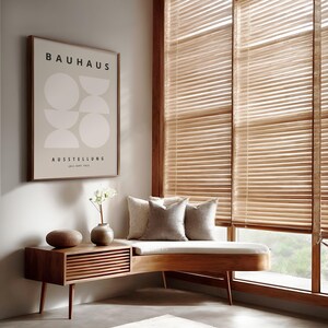 May include: A room corner featuring a wooden bench with two gray pillows, a small wooden side table with a vase and decorative object, and a framed Bauhaus poster. The window has wooden blinds.