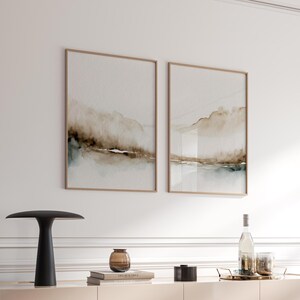 May include: Two framed abstract landscape paintings with brown and green watercolor washes. The paintings are hanging on a white wall above a black table lamp, a brown glass vase, and a stack of books.