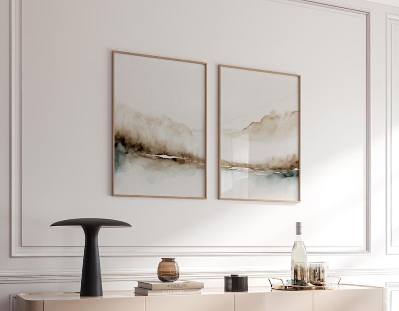 May include: Two framed abstract landscape paintings with brown and green watercolor washes. The paintings are hanging on a white wall above a black table lamp, a brown glass vase, and a stack of books.