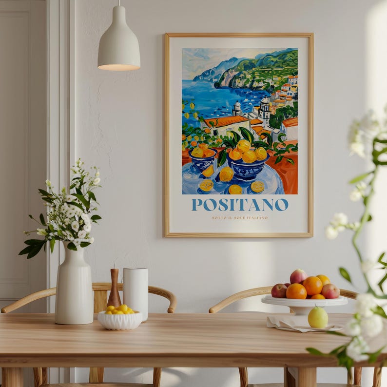 May include: A colorful painting of a coastal town with a bowl of lemons in the foreground. The painting is framed in a light wood frame and has the text "Positano" in blue letters at the bottom. The painting is hanging on a white wall.