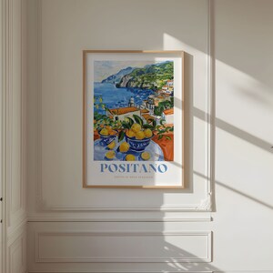 May include: A framed poster with a colorful painting of a coastal town with a blue sea and a mountain in the background. The painting also features a still life of lemons in blue bowls on a table. The poster has the text "POSITANO" in blue letters.