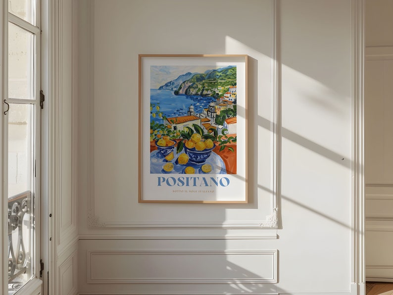 May include: A framed poster with a colorful painting of a coastal town with a blue sea and a mountain in the background. The painting also features a still life of lemons in blue bowls on a table. The poster has the text "POSITANO" in blue letters.