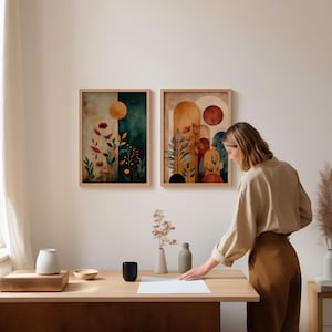 Puede incluir: Dos impresiones art&iacute;sticas en acuarela enmarcadas con temas bot&aacute;nicos. Una presenta flores rojas y un sol amarillo sobre un fondo verde azulado y marr&oacute;n. La otra tiene formas abstractas y plantas en tonos c&aacute;lidos. Las impresiones se exhiben en una pared blanca.