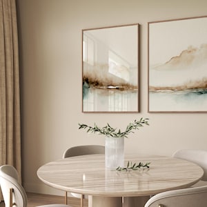 May include: Two framed abstract watercolor paintings with brown and blue hues, hanging on a beige wall above a round wooden table with a vase of greenery in the center.