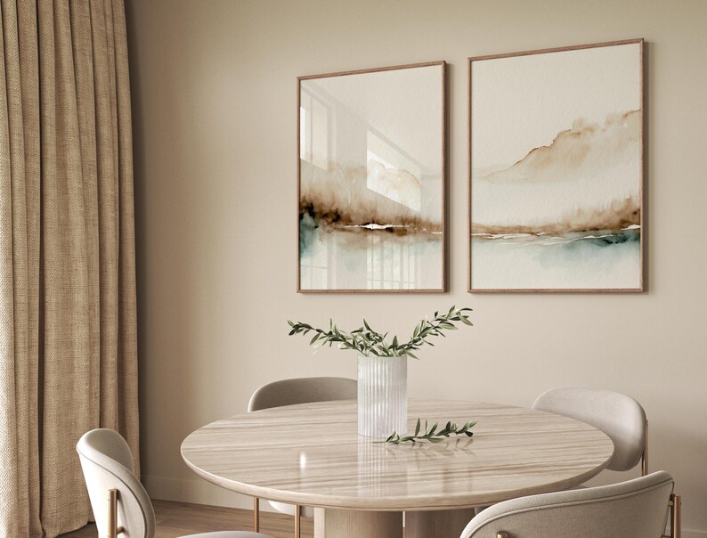 May include: Two framed abstract watercolor paintings with brown and blue hues, hanging on a beige wall above a round wooden table with a vase of greenery in the center.