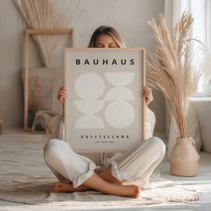 May include: A framed Bauhaus art print featuring a minimalist design of overlapping white shapes on a light gray background. The print includes the word "BAUHAUS" at the top and "AUSSTELLUNG" below, with dates. The artwork is held by a person.