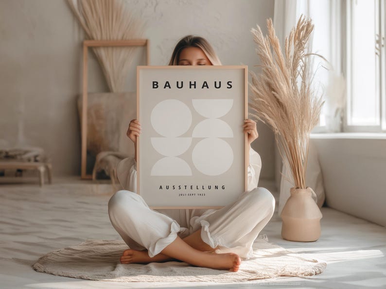 May include: A framed Bauhaus art print featuring a minimalist design of overlapping white shapes on a light gray background. The print includes the word "BAUHAUS" at the top and "AUSSTELLUNG" below, with dates. The artwork is held by a person.