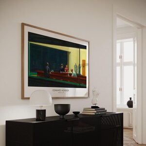 K&ouml;nnte beinhalten: Ein gerahmter Druck von Edward Hoppers Gem&auml;lde "Nighthawks", das eine Diner-Szene mit drei Kunden und einem Kellner zeigt. Der Druck h&auml;ngt an einer wei&szlig;en Wand &uuml;ber einer schwarzen Kommode mit einer Lampe, einer Schale und B&uuml;chern.