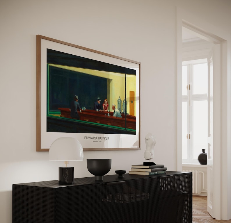 K&ouml;nnte beinhalten: Ein gerahmter Druck von Edward Hoppers Gem&auml;lde "Nighthawks", das eine Diner-Szene mit drei Kunden und einem Kellner zeigt. Der Druck h&auml;ngt an einer wei&szlig;en Wand &uuml;ber einer schwarzen Kommode mit einer Lampe, einer Schale und B&uuml;chern.