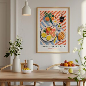 May include: A colorful illustration of a plate of fruit, a croissant, and a glass of juice on a striped tablecloth. The illustration is framed in a light wood frame and has the text "FOOD LOVER CLUB FOOD MARKET" at the bottom.