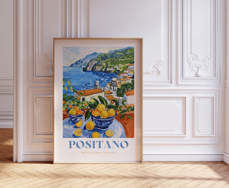 May include: A colorful painting of Positano, Italy, with a blue sky, a turquoise sea, and a hillside town with white buildings. The painting is framed in a gold frame and has the text "Positano Sotto il Sole Italiano" at the bottom.