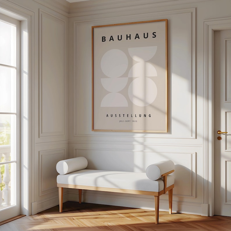 May include: A framed Bauhaus exhibition poster with geometric shapes and text hangs above a white bench with cylindrical bolsters. The bench has a light wood frame. The room has white paneling and a door with a gold handle.
