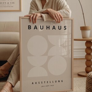 May include: A framed Bauhaus exhibition poster. The poster features a beige background with white geometric shapes and the word "BAUHAUS" in black. Below, the text "AUSSTELLUNG" and "JULI-SEPT 1923" are also in black. The frame is light brown.